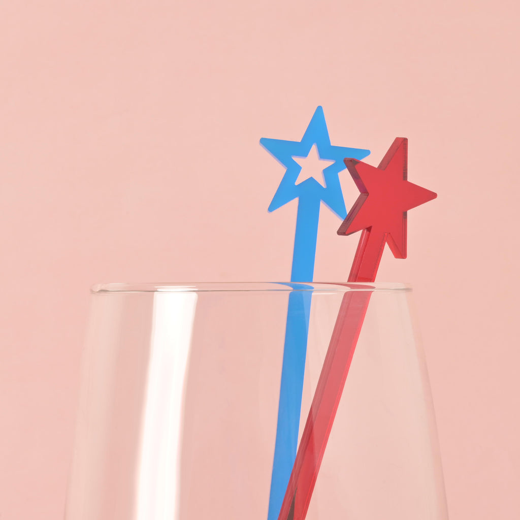 A red and a blue star-shaped cocktail stirrer are placed inside a clear glass. The background is a solid pink color.