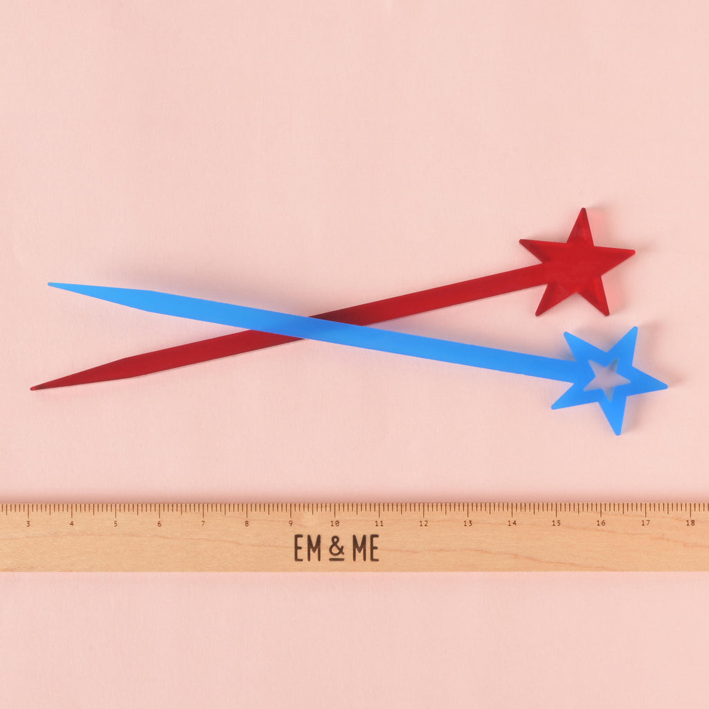 A pair of cocktail stirrers—one red and one blue, both with a star at the top—cross over each other. A ruler with the "EM & ME" logo is placed below the stirrers to show their size. The background is a solid pink color.