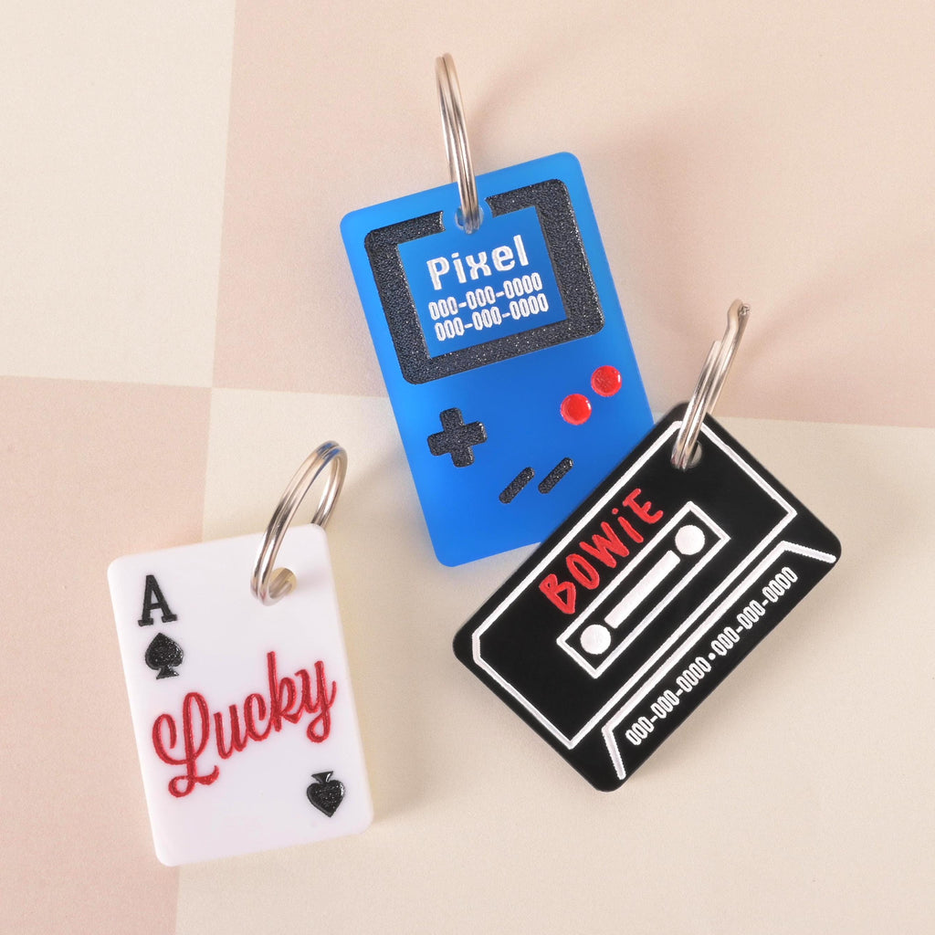 Set of retro pet ID tags including a white playing card with “Lucky,” a blue game console with “Pixel,” and a black cassette tape with “Bowie,” customizable name charms for dogs and cats.