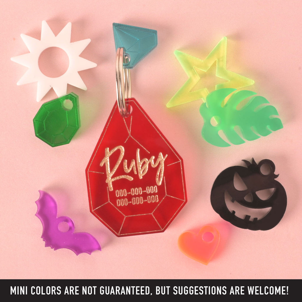 A collection of pet tags and charms scattered on a solid pink background. The central piece is a personalized red gemstone-shaped pet tag with the name "Ruby" and a placeholder phone number on it. A translucent blue diamond charm is attached. Surrounding it are various charms, including a white starburst, a green gemstone, a neon yellow star, a green monstera leaf, a purple bat, an orange heart, and a black carved pumpkin. The image also contains the text "MINI COLORS ARE NOT GUARANTEED, BUT SUGGESTIONS ARE