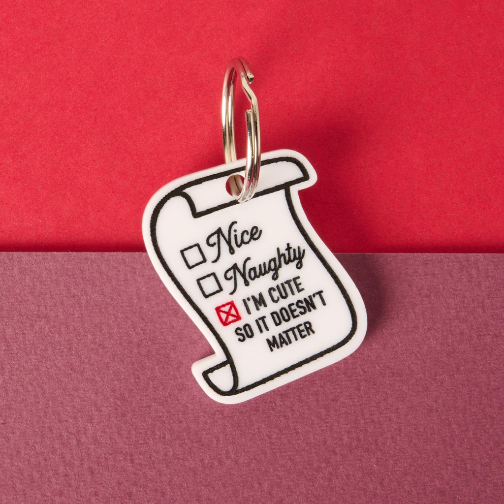 A white scroll-shaped pet tag featuring a naughty or nice checklist. The tag is marked with a red 'X' next to the text "I'M CUTE SO IT DOESN'T MATTER." The tag is attached to a silver ring, perfect for a collar, and is displayed on a background split between solid red and solid purple.