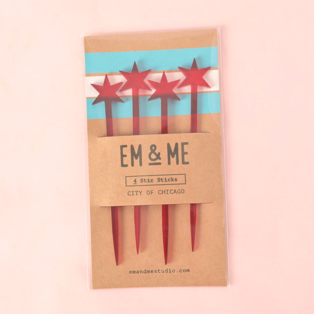 A packaged set of four red cocktail stirrers, each topped with a star resembling the Chicago flag. The packaging is a brown card that features the flag's blue and white stripes and the text "EM & ME" and "CITY OF CHICAGO." The item is displayed on a solid pink background.