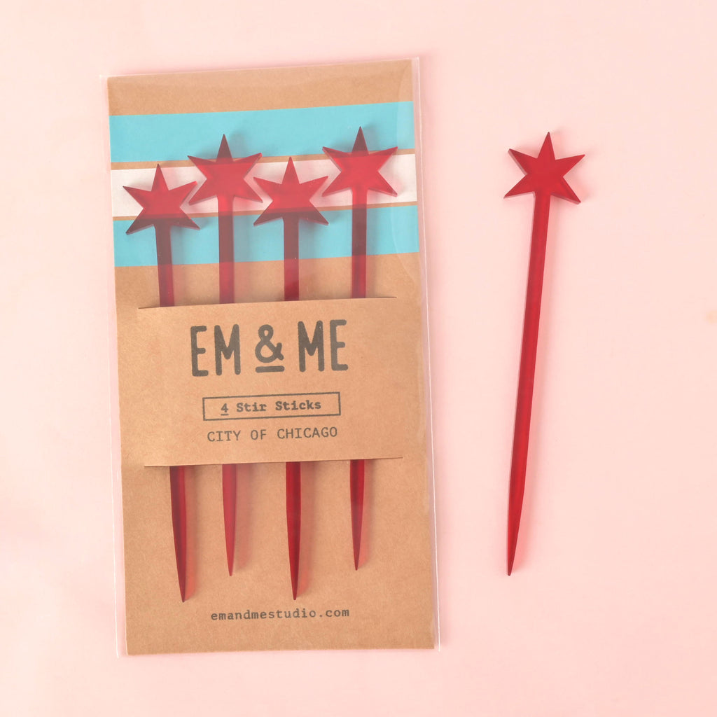 A packaged set of four red cocktail stirrers, each topped with a star resembling the Chicago flag. The packaging is a brown card that features the flag's colors and the text "EM & ME" and "CITY OF CHICAGO." One matching red star stirrer is shown next to the package. The items are displayed on a solid pink background.