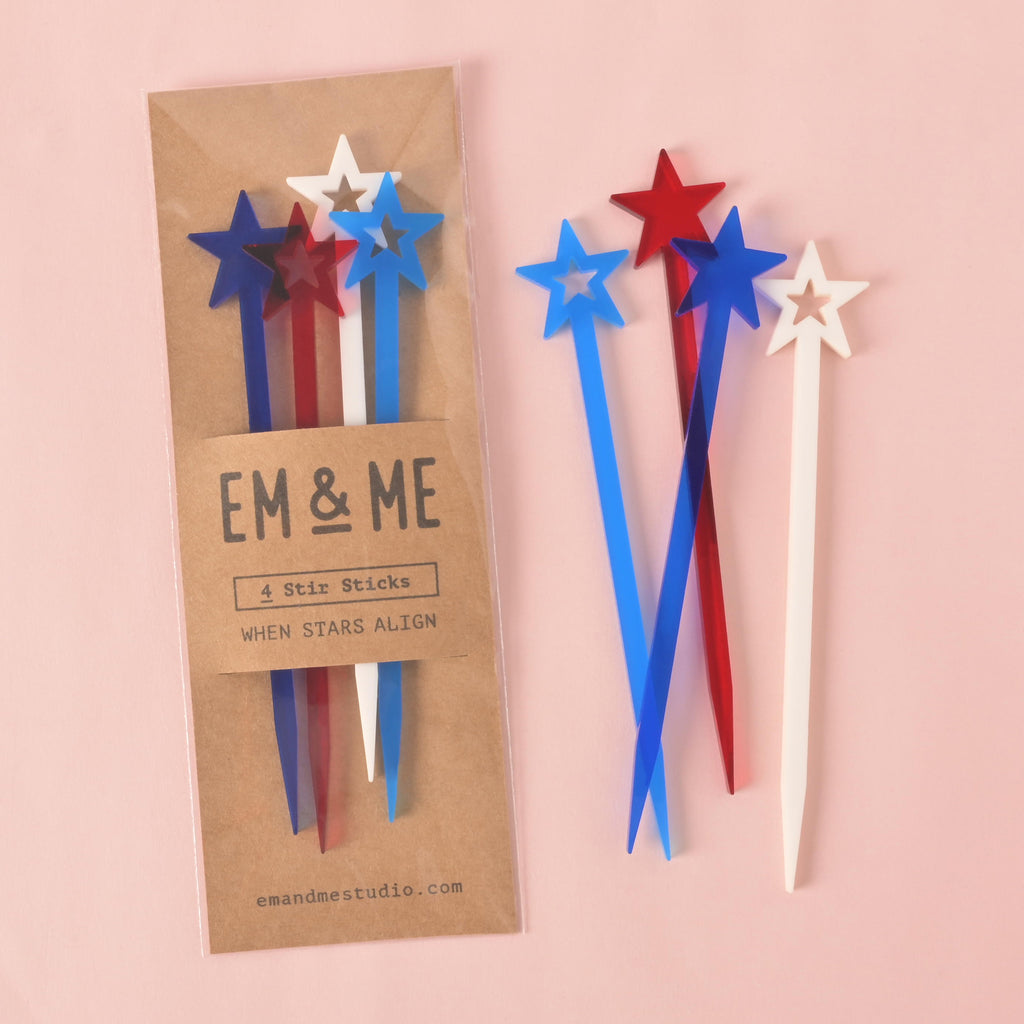 A packaged set of four red, white, and blue star-shaped cocktail stirrers. A second set of three stirrers—a white, blue, and red—is shown outside the package. The brown cardstock packaging has "EM & ME" and "When Stars Align" printed on it. The background is a solid pink color.