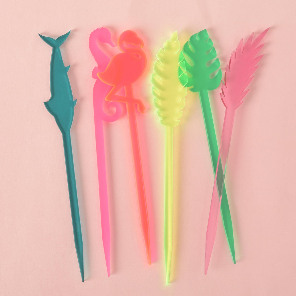 A collection of six assorted tropical cocktail stirrers on a solid pink background. The stirrers include a teal swordfish, a fluorescent pink flamingo, a fluorescent green palm frond, a frosted pink seahorse, a green monstera leaf, and a transparent pink palm frond.