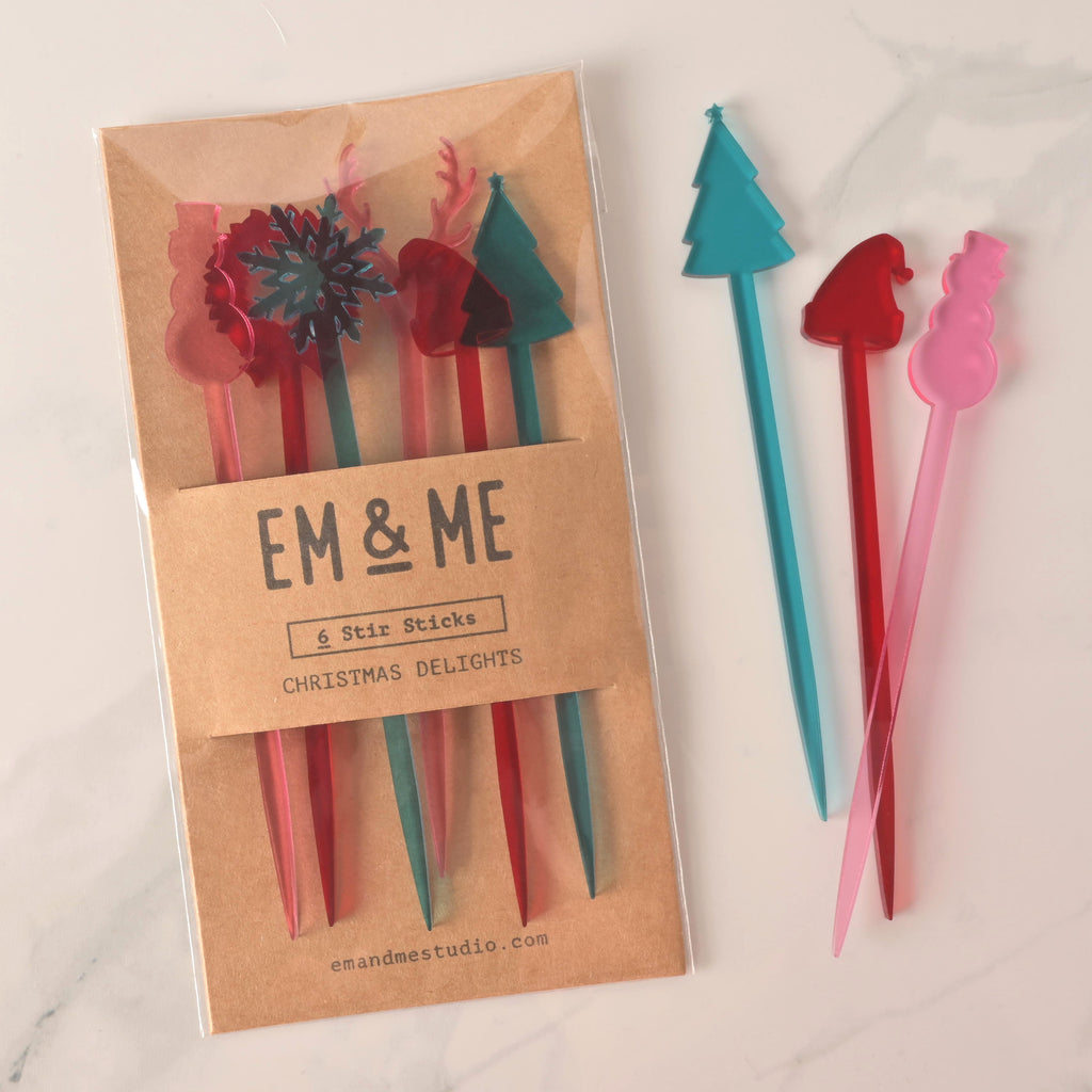 A packaged set of six colorful "Christmas Delights" cocktail stirrers in red, pink, and teal. The package includes various shapes like a snowflake, reindeer, and Christmas tree. Three loose stirrers—a teal Christmas tree, a red Santa hat, and a pink snowman—are shown next to the package. The brown card packaging reads "EM & ME" and "6 Stir Sticks." The items are displayed on a white marble surface.