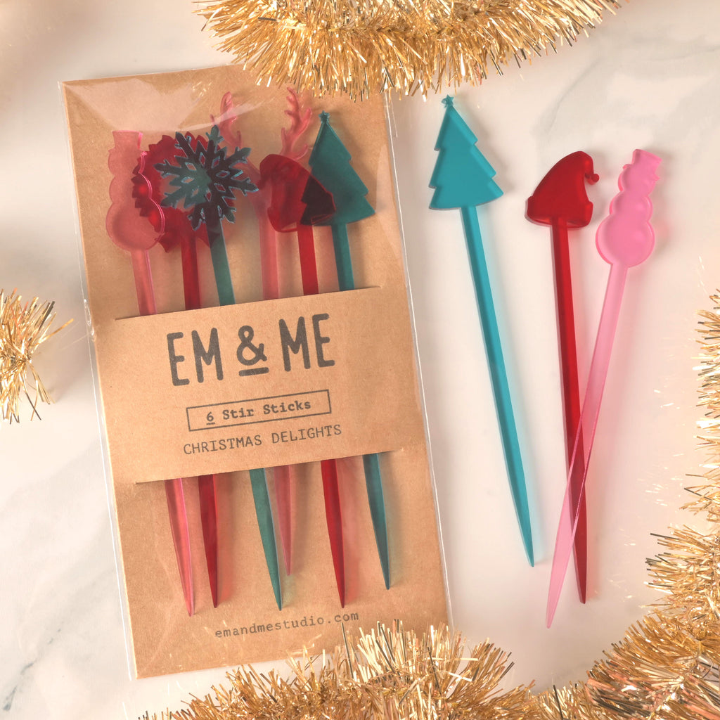 A packaged set of six colorful "Christmas Delights" cocktail stirrers in red, pink, and teal. The package includes various shapes like a snowflake, reindeer, and Christmas tree. Three loose stirrers—a teal Christmas tree, a red Santa hat, and a pink snowman—are shown next to the package. The brown card packaging reads "EM & ME" and "6 Stir Sticks." The items are displayed on a white marble surface.
