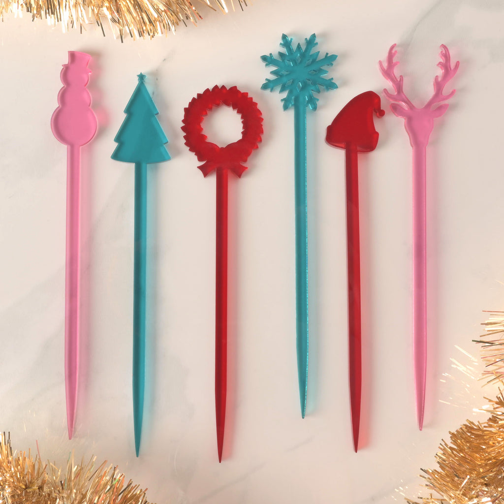 A set of six colorful Christmas-themed cocktail stirrers, arranged side-by-side on a white marble surface with golden tinsel. The pink stirrers are shaped like a snowman and a reindeer, the blue stirrers are a Christmas tree and a snowflake, and the red stirrers are a wreath and a Santa hat.