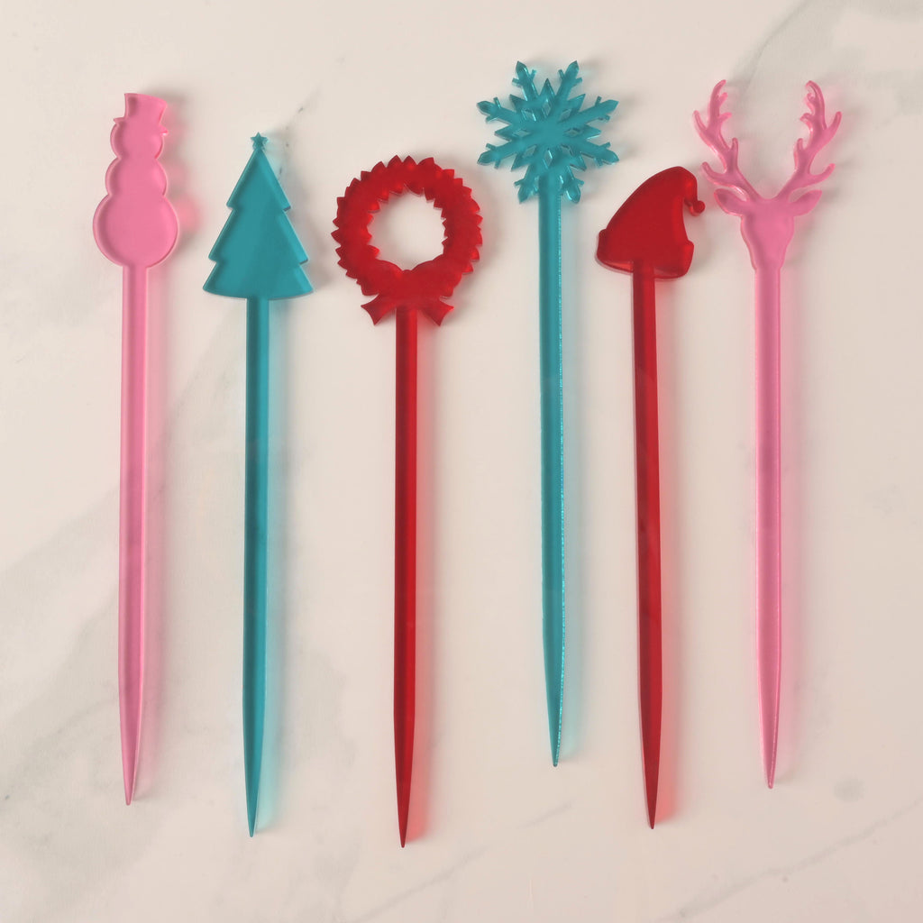 A set of six colorful Christmas-themed cocktail stirrers, arranged side-by-side on a white marble surface. The stirrers include a pink snowman, a blue Christmas tree, a red wreath, a blue snowflake, a red Santa hat, and a pink reindeer (or buck head).