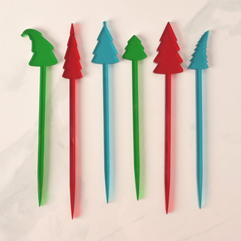 A set of six Christmas tree-shaped cocktail stirrers in red, green, and blue. The stirrers feature slightly different tree shapes and heights. They are arranged side-by-side on a white marble surface.