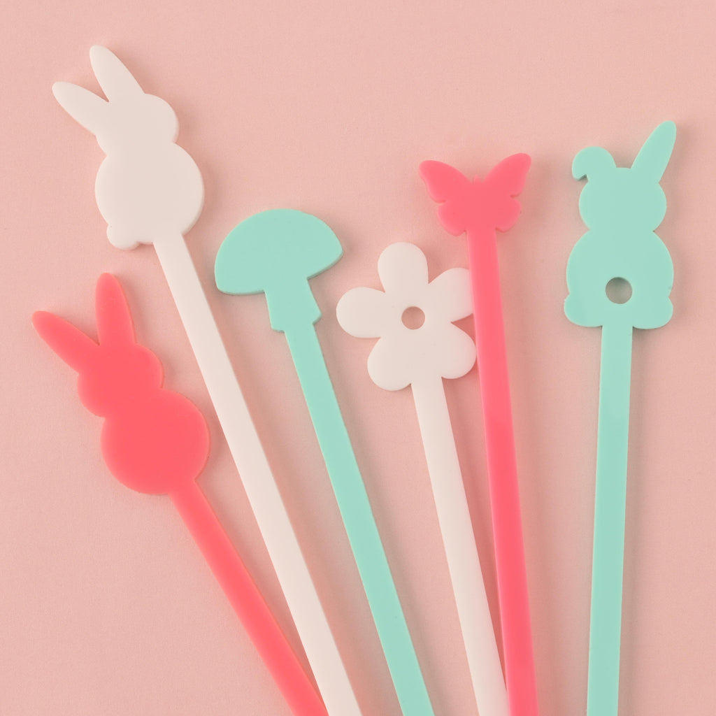 A set of six colorful cocktail stirrers, including two pink bunnies, a light blue mushroom, a white flower, a pink butterfly, and a light blue bunny. The items are arranged in a scattered group on a solid pink background.