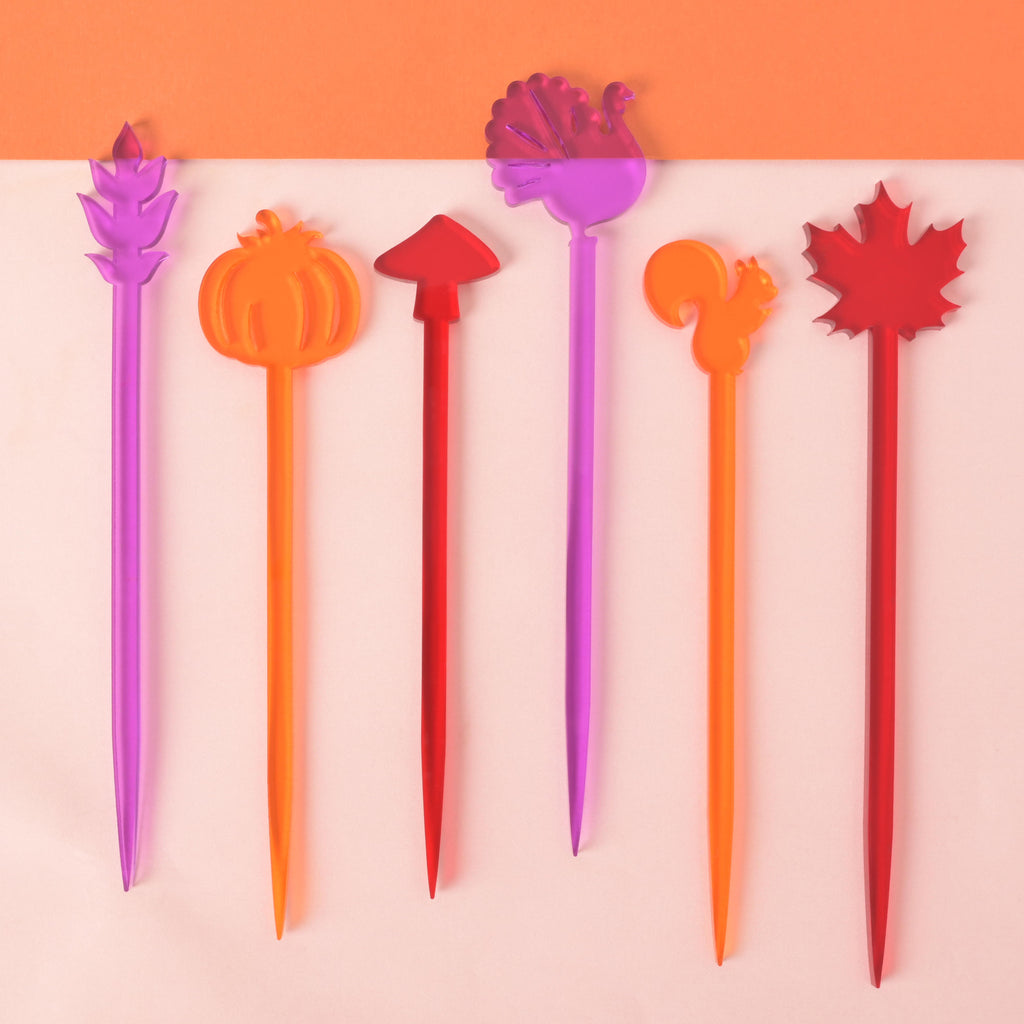A set of six colorful cocktail stirrers in purple, orange, and red, featuring various autumn and Thanksgiving shapes. The shapes include a wheat stalk, a pumpkin, a mushroom, a turkey, a squirrel, and a maple leaf. The items are displayed on a solid light pink and orange background.