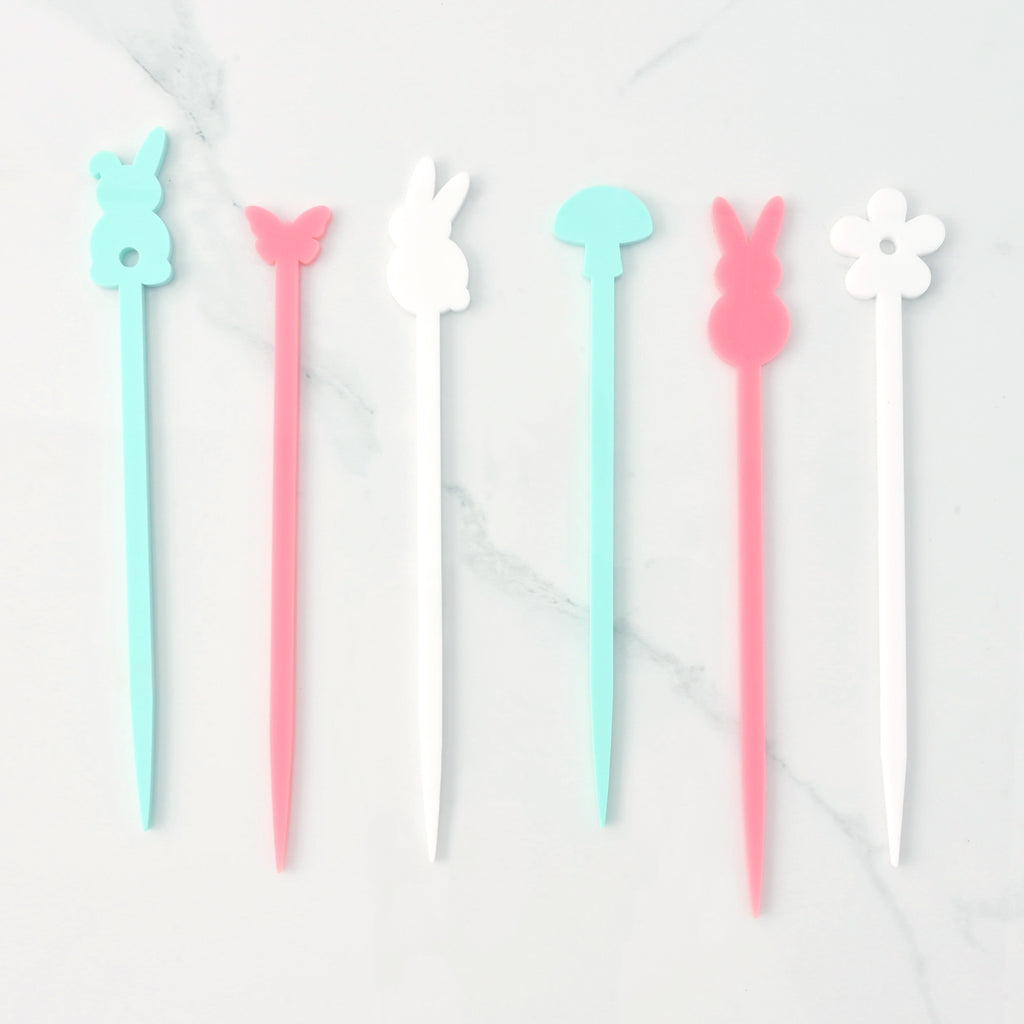  set of six colorful cocktail stirrers. From left to right, the stirrers are a light blue bunny, a pink butterfly, a white bunny, a light blue mushroom, a pink bunny, and a white flower. The items are shown on a light-colored, speckled surface.