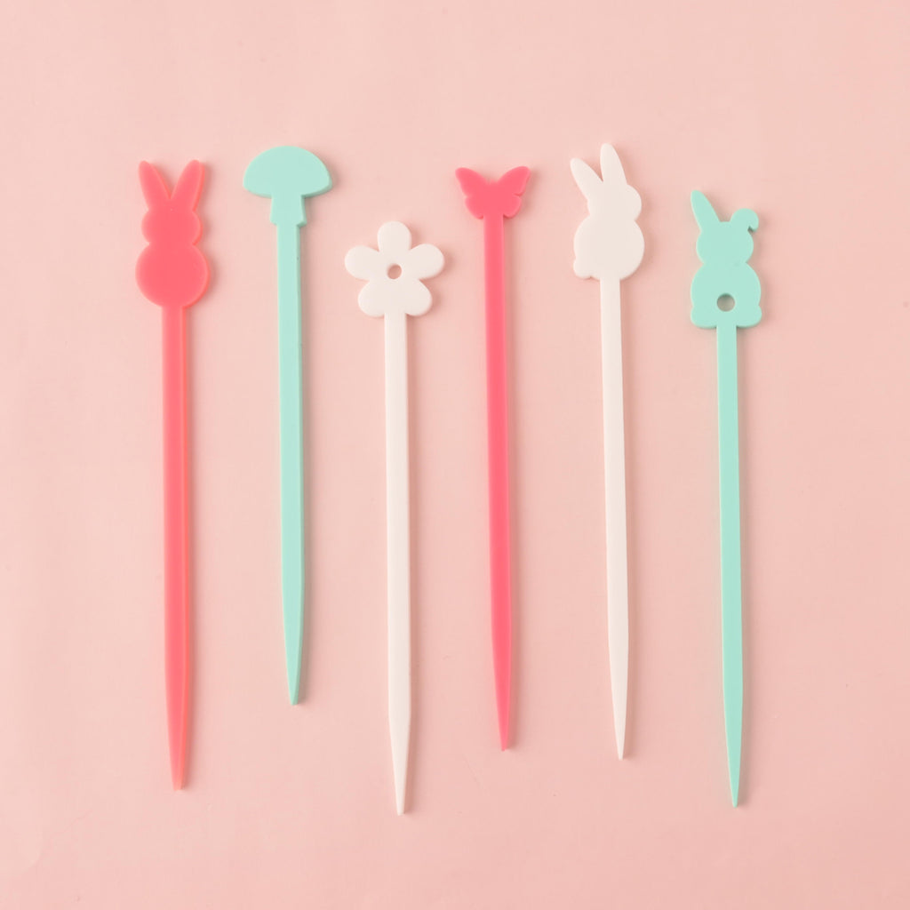 A set of six colorful cocktail stirrers. From left to right, the stirrers are a pink bunny, a light blue mushroom, a white flower, a pink butterfly, a white bunny, and a light blue bunny. The items are shown on a solid pink background.