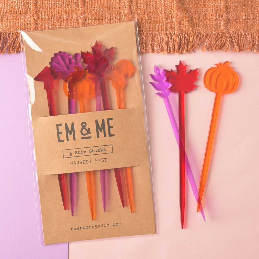 A packaged set of six "Harvest Fest" cocktail stirrers in autumnal colors like purple, red, and orange. The stirrers are shaped like a maple leaf, a pumpkin, and other fall icons. Three loose stirrers are shown next to the package: a purple leaf, a red maple leaf, and an orange pumpkin. The package reads "EM & ME" and "6 Stir Sticks." The items are displayed on a background with a purple and pink surface and an orange knitted blanket.