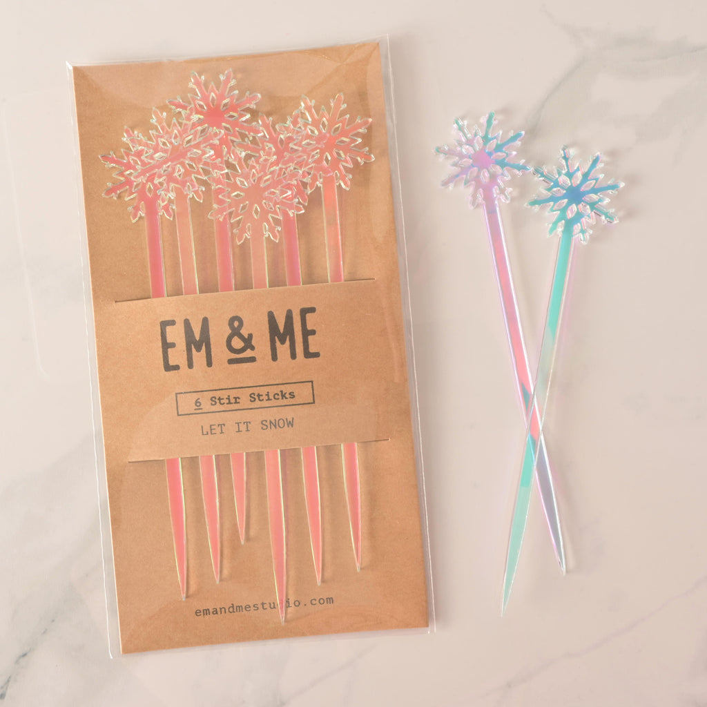 A packaged set of six pink and blue iridescent snowflake-topped cocktail stirrers. Two loose, iridescent snowflake stirrers are shown crossed next to the package. The brown card packaging reads "EM & ME" and "6 Stir Sticks" with the theme "LET IT SNOW." The items are displayed on a white marble surface.