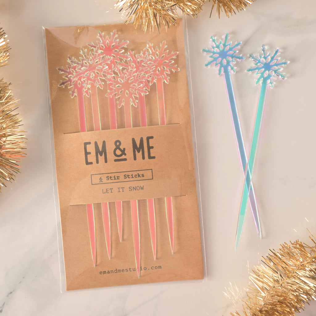 A packaged set of six pink and blue iridescent snowflake-topped cocktail stirrers. Two loose, blue and pink iridescent snowflake stirrers are shown next to the package. The brown card packaging reads "EM & ME" and "6 Stir Sticks" with the theme "LET IT SNOW." The items are displayed on a white marble surface with golden tinsel decorations.