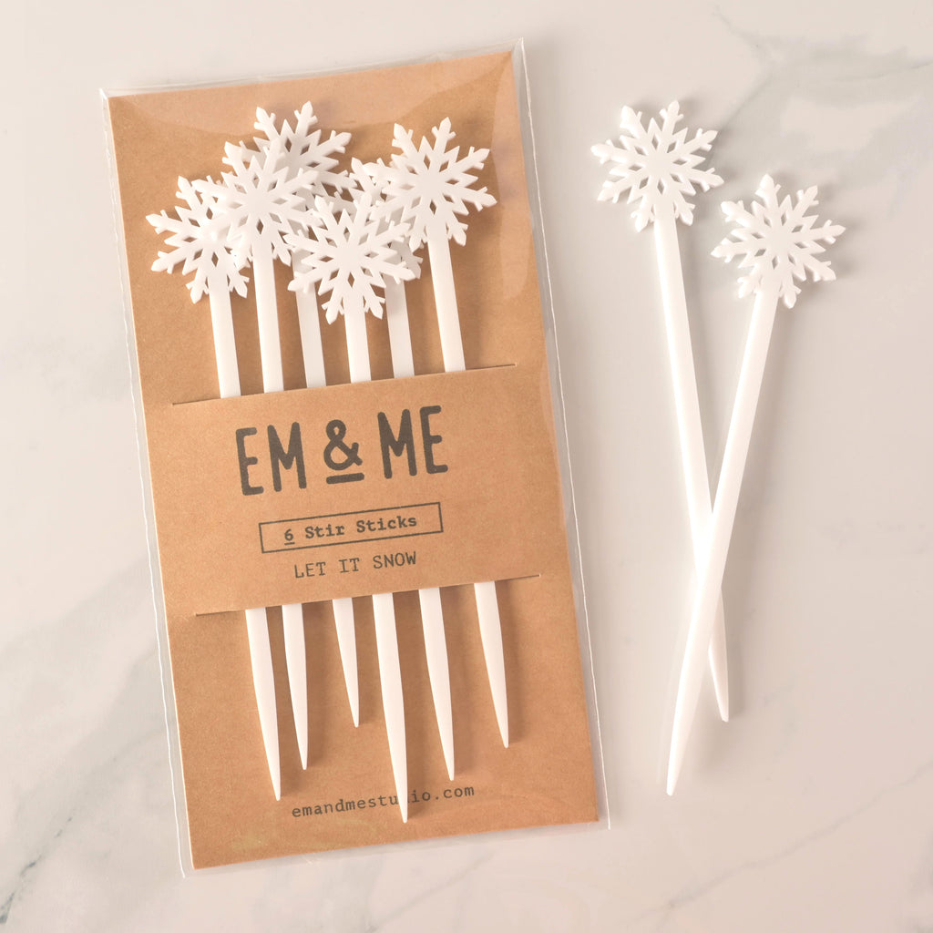 A packaged set of six white snowflake-topped cocktail stirrers. Two loose stirrers are shown next to the package. The brown card packaging reads "EM & ME" and "6 Stir Sticks" with the theme "LET IT SNOW." The items are displayed on a white marble surface.