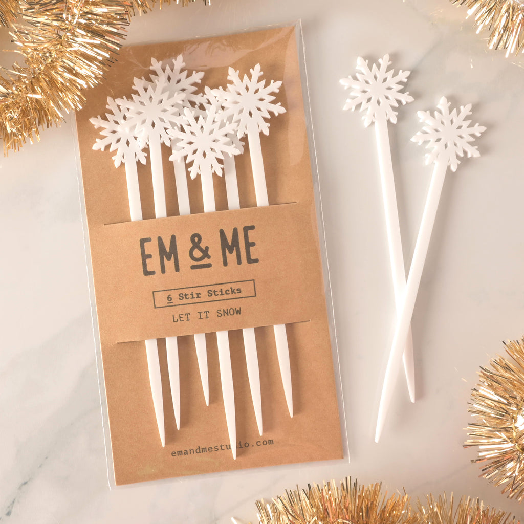 A packaged set of six white snowflake-topped cocktail stirrers. Two loose stirrers are shown next to the package. The brown card packaging reads "EM & ME" and "6 Stir Sticks" with the theme "LET IT SNOW." The items are displayed on a white marble surface with golden tinsel decorations.
