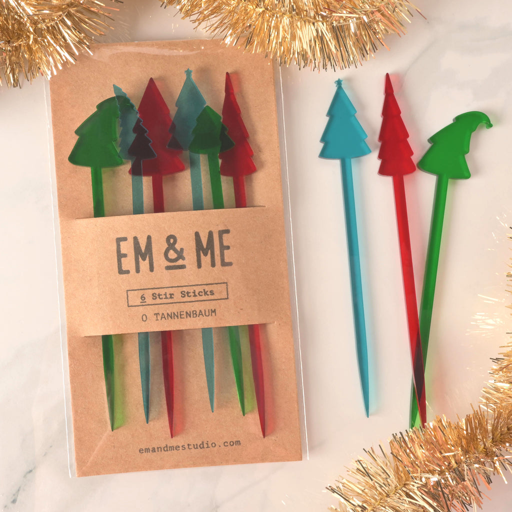 A packaged set of six "O Tannenbaum" Christmas tree-shaped cocktail stirrers in red, green, and blue. Three loose stirrers—a blue, red, and green tree—are shown next to the package. The brown card packaging reads "EM & ME" and "6 Stir Sticks." The items are displayed on a white marble surface with golden tinsel decorations.