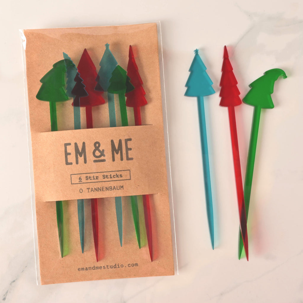 A packaged set of six "O Tannenbaum" Christmas tree-shaped cocktail stirrers in red, green, and blue. Three loose stirrers—a blue, red, and green tree—are shown next to the package. The brown card packaging reads "EM & ME" and "6 Stir Sticks." The items are displayed on a white marble surface.
