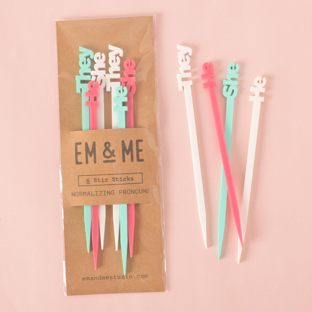 A packaged set of six pronoun cocktail stirrers, including two for "he," two for "she," and two for "they." The stirrers are in pink, white, and light blue. Two stirrers are shown outside the package. The brown cardstock packaging has "EM & ME" and "Normalizing Pronouns" printed on it. The background is a solid pink color.