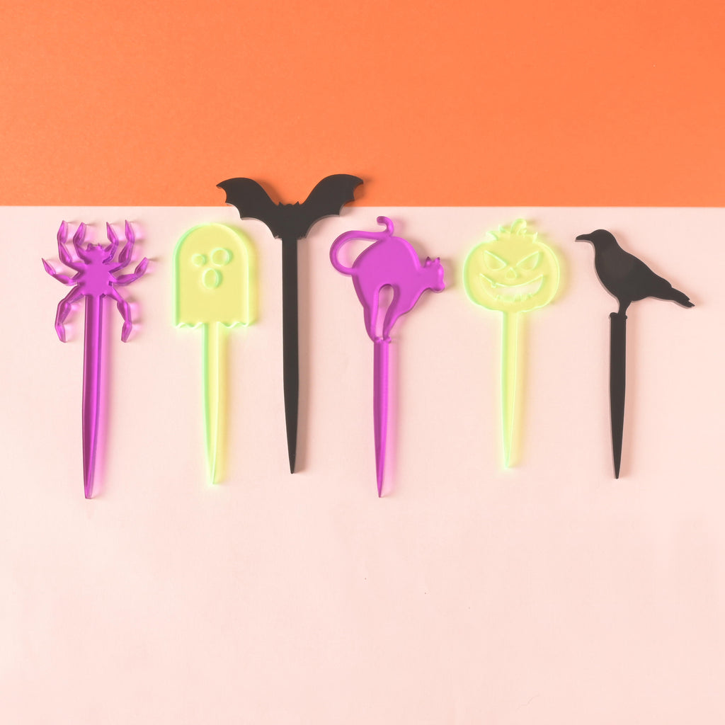Collection of 6 halloween themed food picks. Purple spider, fluorescent green ghost, black bat, purple cat, fluorescent green jack-o-lantern and black crown on color blocked background