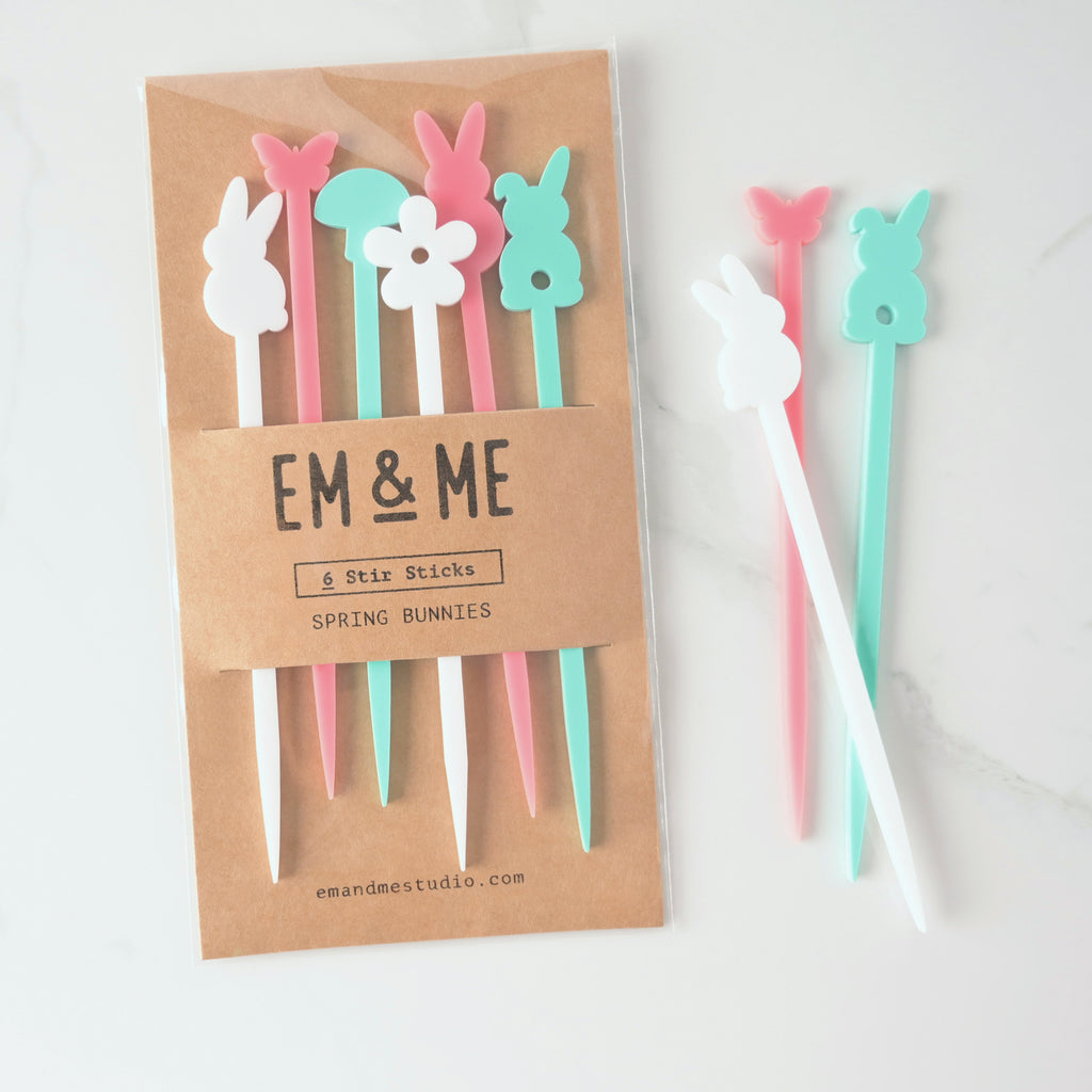 A packaged set of six "Spring Bunnies" stirrers in various colors, including a white bunny, a pink butterfly, a light blue bunny, a pink bunny, and a white flower. Three of the stirrers—a white bunny, a pink butterfly, and a light blue bunny—are shown outside the package. The brown cardstock packaging has "EM & ME" and "Spring Bunnies" printed on it. The items are shown on a white marble surface.