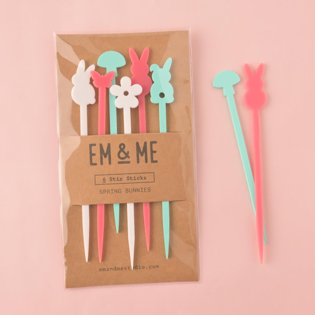A packaged set of six "Spring Bunnies" stirrers in various colors, including a white bunny, a pink bunny, a light blue bunny, a pink butterfly, and a white flower. Two of the stirrers—a light blue mushroom and a pink bunny—are shown outside the package. The brown cardstock packaging has "EM & ME" and "Spring Bunnies" printed on it. The items are shown on a solid pink background.