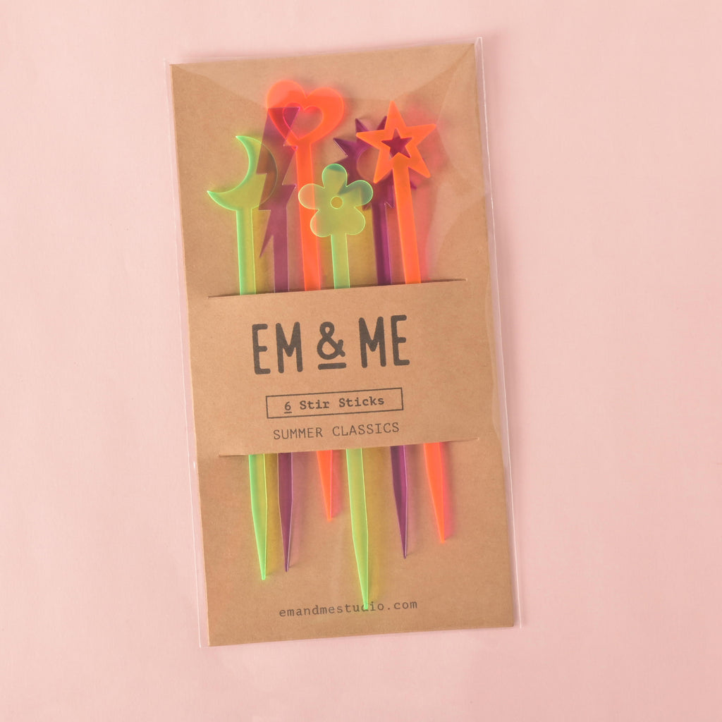 A packaged set of six colorful "Summer Classics" cocktail stirrers, including a moon, a heart, a star, and a flower. The stirrers are in Fluorescent pink, green, and purple. The brown cardstock packaging has "EM & ME" and "Summer Classics" printed on it. The background is a solid pink color.