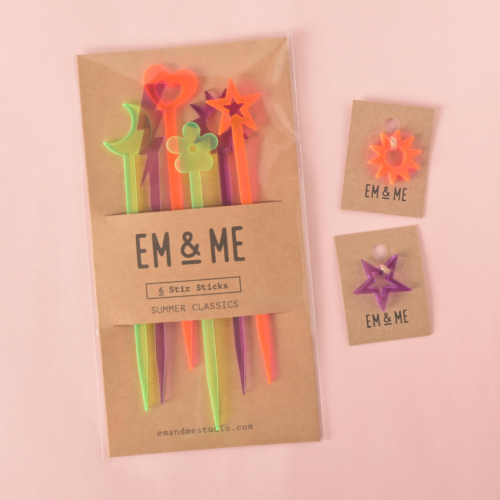 A packaged set of six colorful "Summer Classics" cocktail stirrers, including a heart, a flower, a star, and other shapes. Also shown are two small star-shaped mini charms in fluorescent pink and purple, each on a brown card. All products are from "EM & ME" and are shown on a solid pink background.