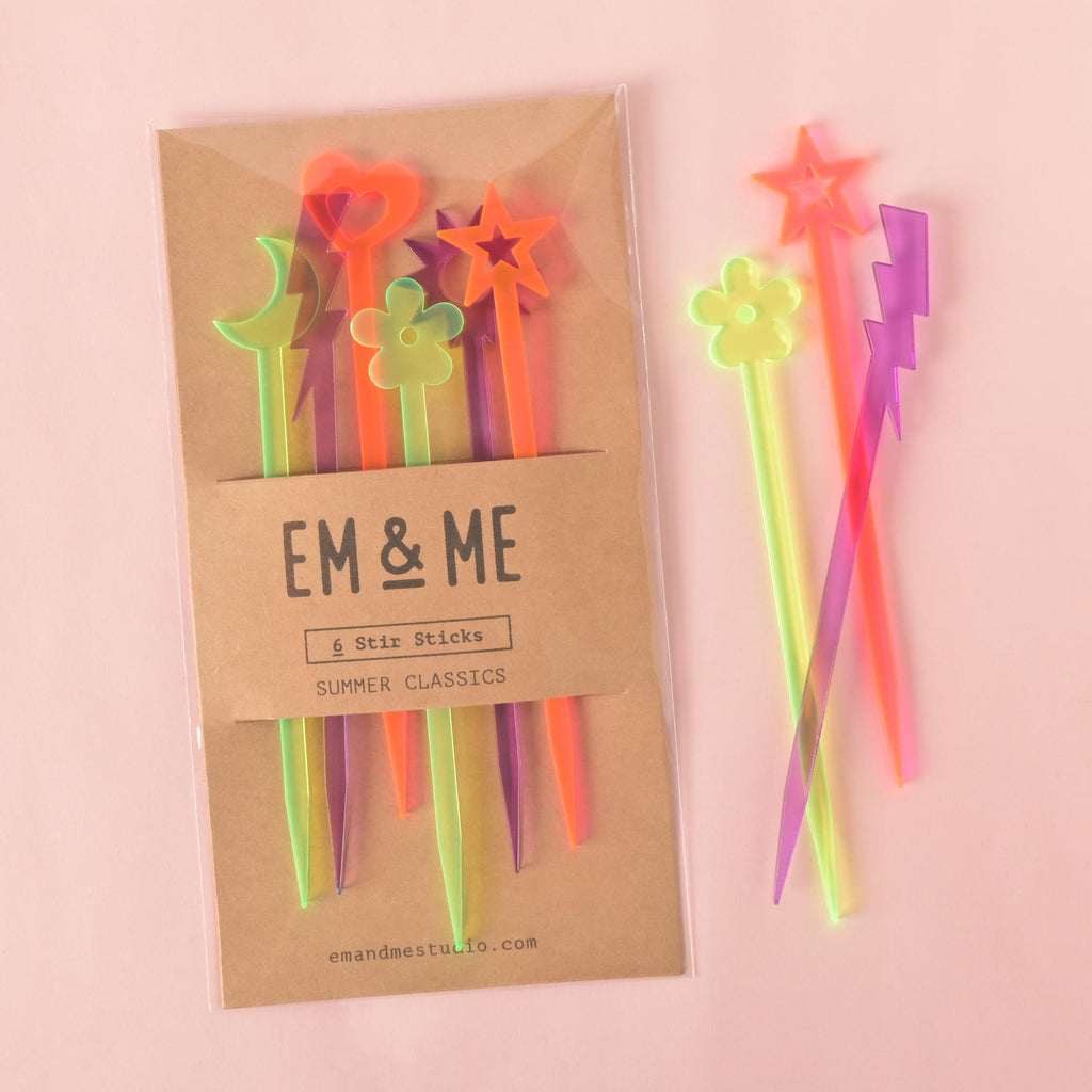 A packaged set of six colorful "Summer Classics" cocktail stirrers, including a moon, a heart, a flower, and a star. Two of the stirrers—a fluorescent green flower and a fluorescent pink star—are shown outside the package, along with a purple lightning bolt stirrer. The brown cardstock packaging has "EM & ME" and "Summer Classics" printed on it. The background is a solid 