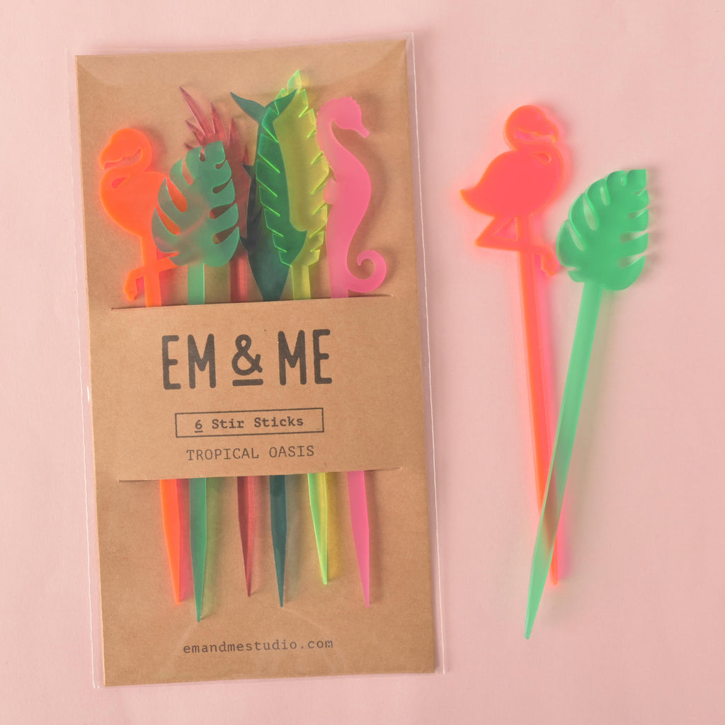 A packaged set of six tropical cocktail stirrers, including a fluorescent pink flamingo, a green monstera leaf, a neon yellow palm frond, a teal dolphin, a pink seahorse, and a neon pink palm frond. Two of the stirrers are shown outside the package. The brown cardstock packaging has "EM & ME" and "Tropical Oasis" printed on it. The background is a solid pink color.