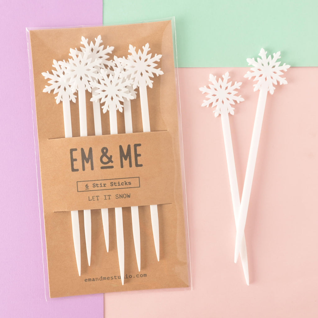 A packaged set of six white snowflake-topped cocktail stirrers. Two loose stirrers are shown crossed next to the package. The brown card packaging reads "EM & ME" and "6 Stir Sticks" with the theme "LET IT SNOW." The items are displayed on a background split between solid pink, light purple, and mint green.