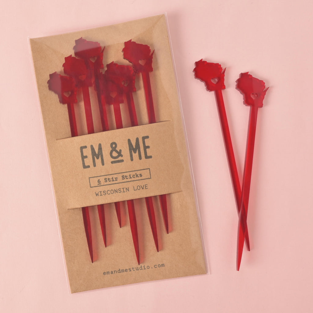 A packaged set of six red cocktail stirrers shaped like the state of Wisconsin. Two stirrers are shown outside the package. The brown cardstock packaging has "EM & ME" and "6 Stir Sticks" printed on it. The background is a solid pink color.