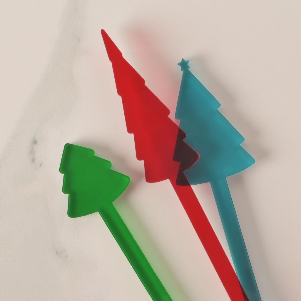 A group of three Christmas tree-shaped cocktail stirrers in green, red, and blue. The stirrers are overlapping on a white marble surface. The blue stirrer is topped with a small star.