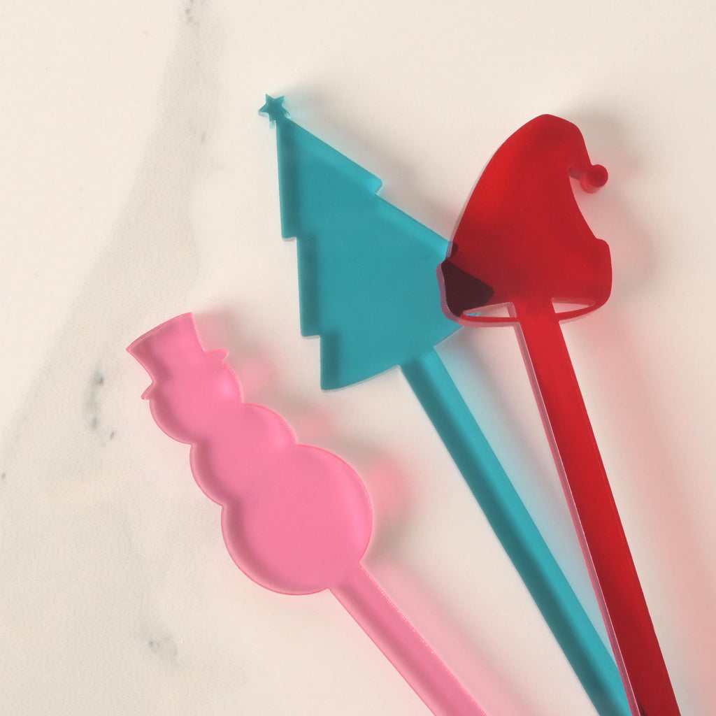 A close-up of three colorful Christmas-themed cocktail stirrers overlapping on a white marble surface. The stirrers are a pink snowman, a blue Christmas tree topped with a star, and a red Santa hat.