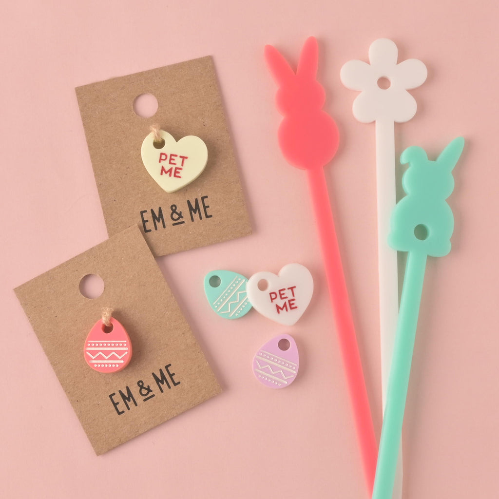 A collection of Easter-themed stirrers and charms. The items include a pink bunny stirrer, a white flower stirrer, a light blue bunny stirrer, a light yellow heart charm on a brown card that says "PET ME," and a pink and white egg charm on a brown card. There are also two small loose charms—a heart and an egg—with a zigzag pattern. The items are shown on a solid pink background.