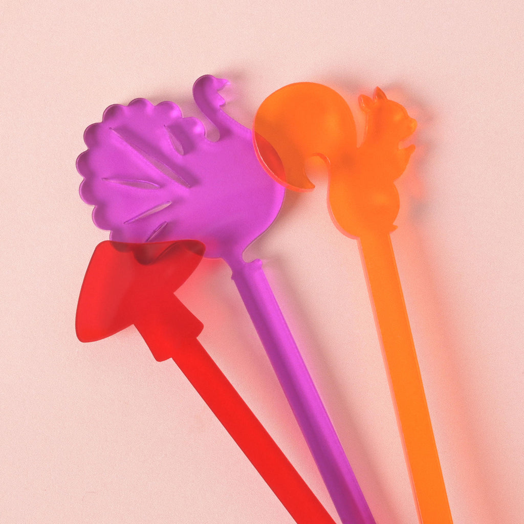 A close-up of four colorful, fall-themed cocktail stirrers overlapping on a solid pink background. The stirrers are shaped like a purple turkey, a bright orange squirrel, a purple mushroom, and a red arrow.