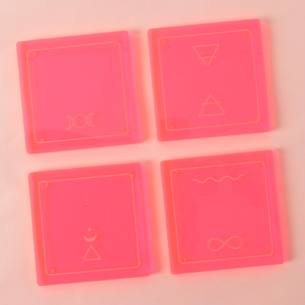 A set of four square, translucent pink coasters arranged on a light pink background. Each coaster has a gold-etched design. The symbols include a pair of triangles, a triple moon, an infinity sign and wave, and a crescent moon with a triangle.