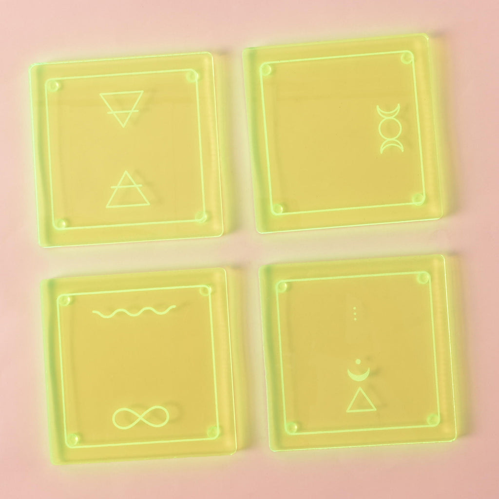 A set of four square, translucent Fluorescent Green coasters arranged in a grid on a light pink background. Each coaster has a unique gold-etched design. The symbols include a pair of triangles, a triple moon, an infinity sign with a wave, and a crescent moon with a triangle.