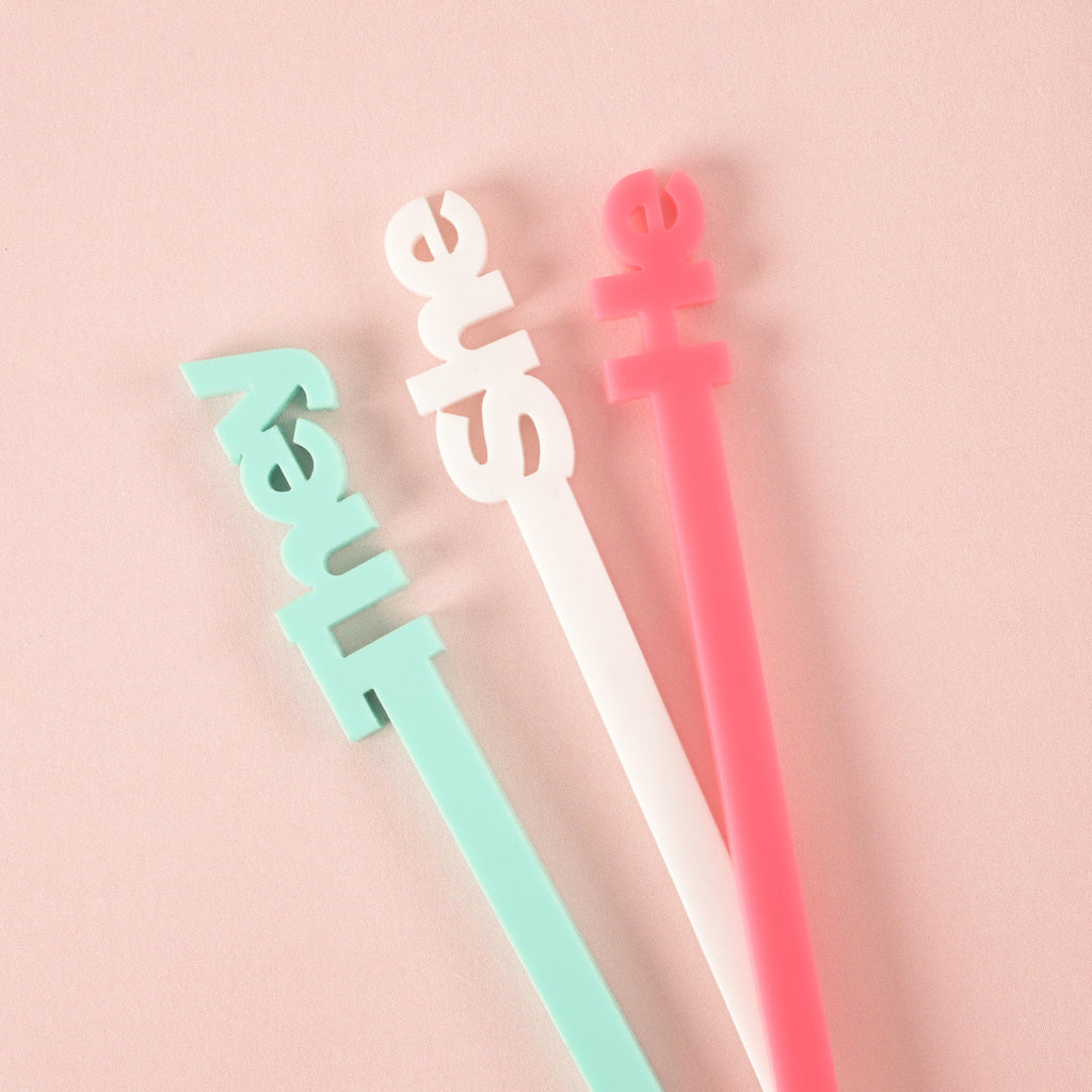 A close-up of three cocktail stirrers with the words "they" in light blue, "she" in white, and "he" in pink, fanned out on a solid pink background.