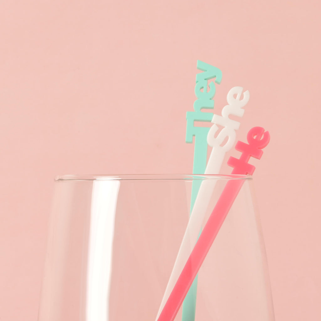 A set of three pronoun cocktail stirrers in different colors—pink for "he," white for "she," and light blue for "they"—placed inside a clear glass. The background is a solid pink color.
