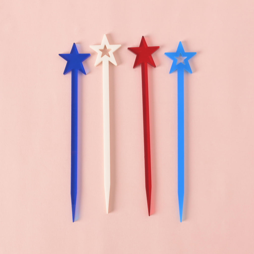 A collection of four red, white, and blue star-shaped cocktail stirrers. The stirrers are positioned in a row on a solid pink background.