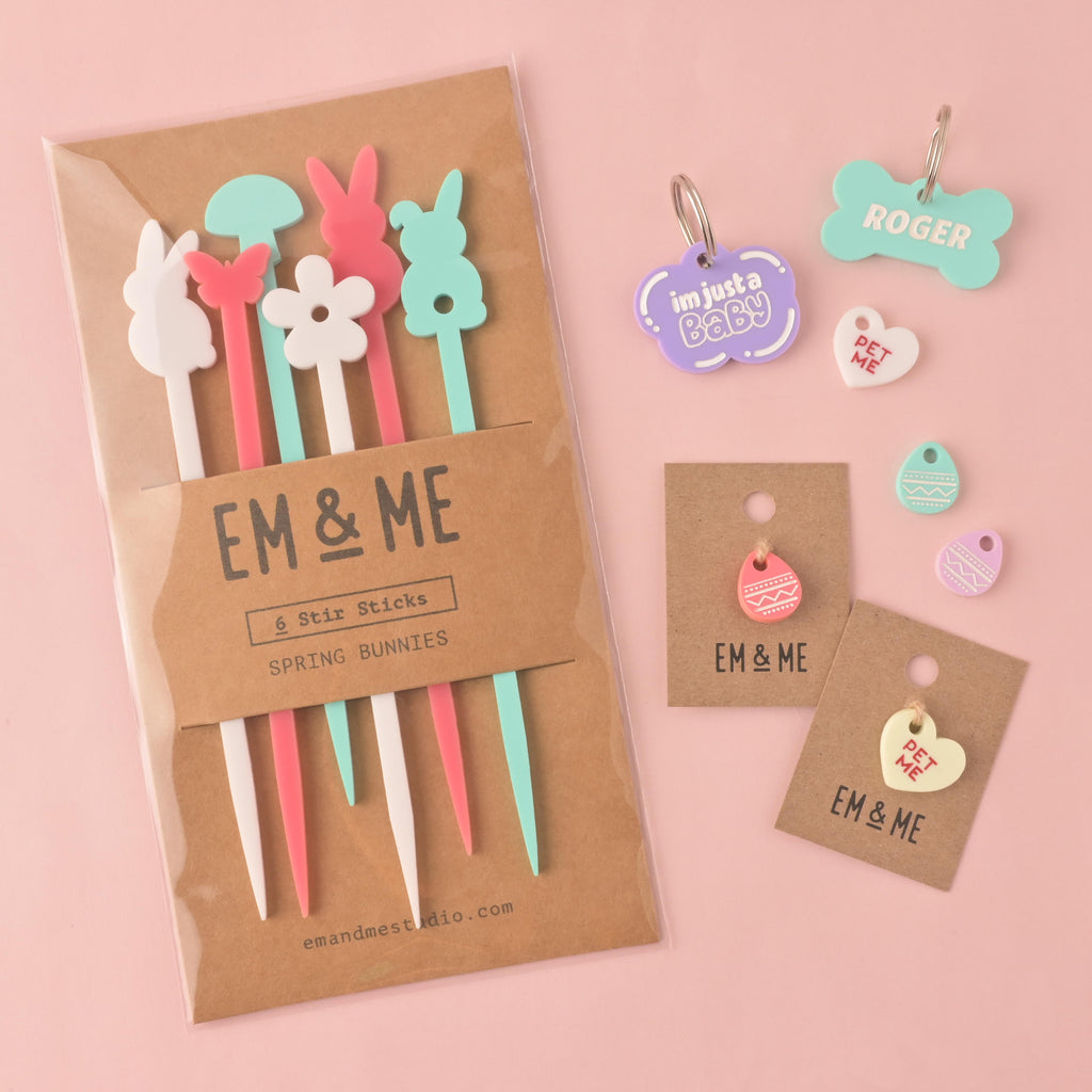 A packaged set of six "Spring Bunnies" stirrers in various colors, including a white bunny, a pink bunny, a white flower, a light blue bunny, and a pink stirrer. Also shown are a purple "I'm just a Baby" cloud-shaped pet tag, a blue bone-shaped tag that says "ROGER," a light yellow "PET ME" heart charm on a brown card, and a pink and white egg charm on a brown card. The items are shown on a solid pink background.