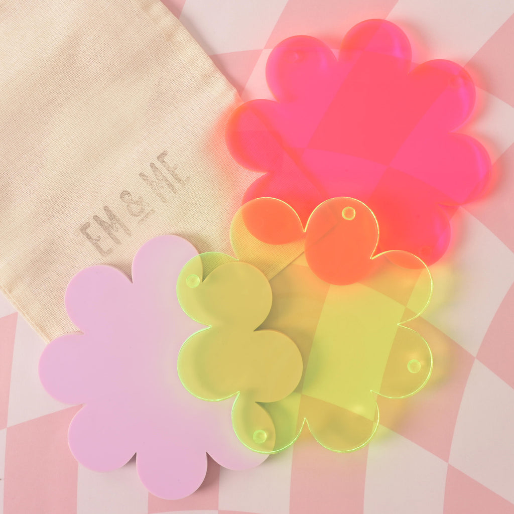 A group of three flower-shaped coasters in different colors—Lavender, translucent Fluorescent Green, and neon pink—is arranged on a pink and white checkered background. A beige pouch with the "EM & ME" logo is in the top-left corner.