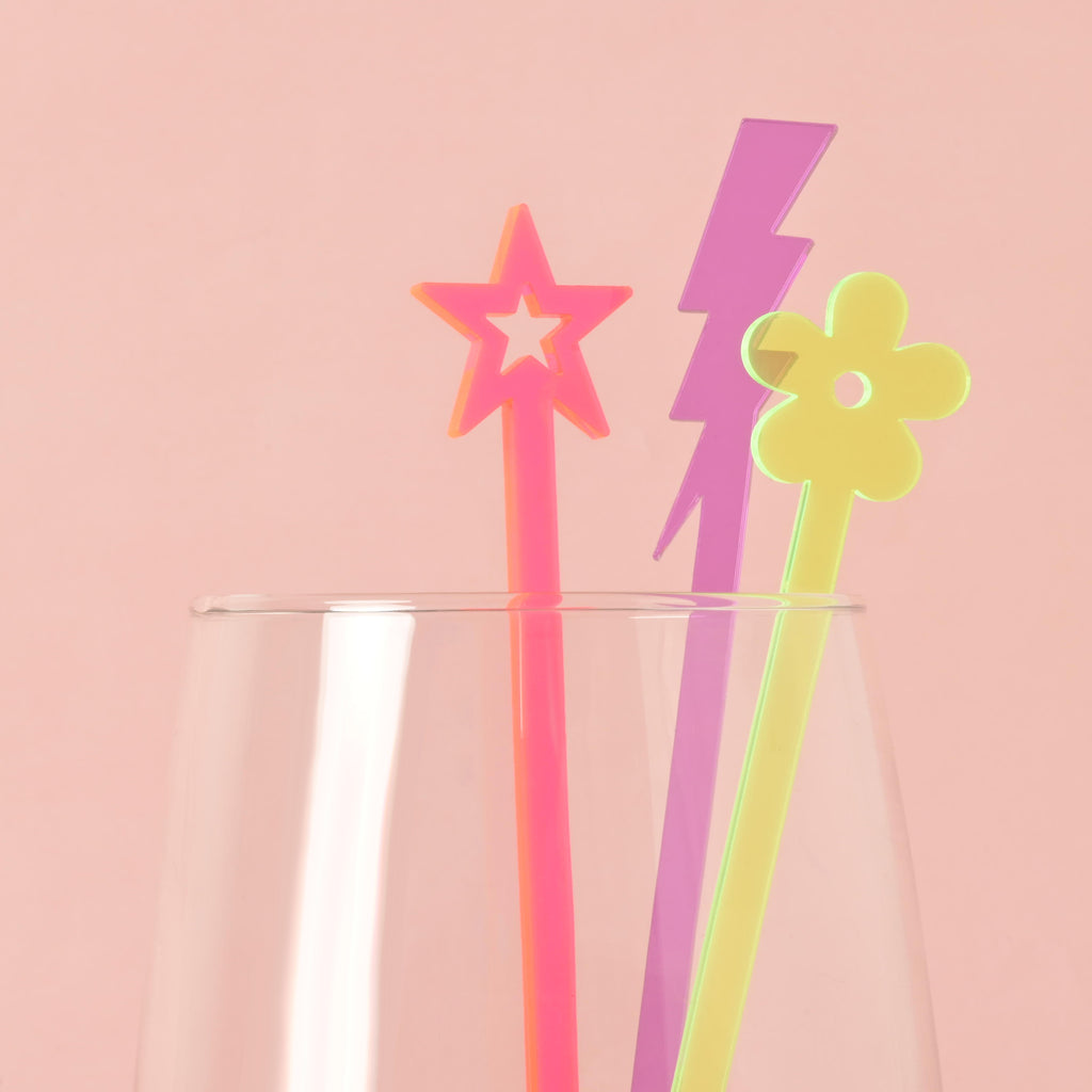 A set of three colorful cocktail stirrers—a neon pink star, a purple lightning bolt, and a yellow flower—are placed inside a clear glass. The background is a solid pink color.