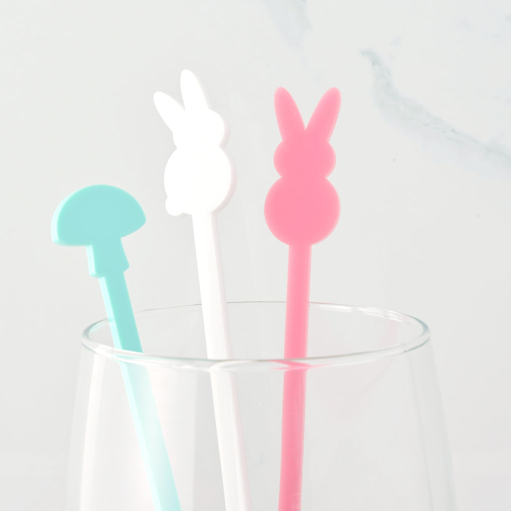 Three Easter-themed cocktail stirrers—a light blue mushroom, a white bunny, and a pink bunny—are standing in a clear glass. The background is a white marble surface.