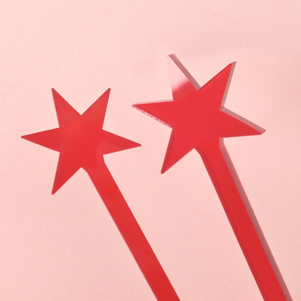 A close-up of two red, transparent cocktail stirrers, each topped with a six-pointed star resembling the Chicago Flag. The stirrers overlap on a solid pink background.