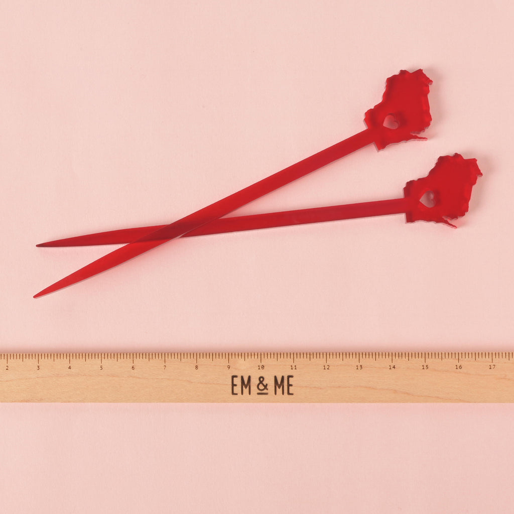 A pair of red cocktail stirrers shaped like the state of Wisconsin, each with a heart cut out in the middle. A ruler with the "EM & ME" logo is placed below the stirrers to show their size. The background is a solid pink color.
